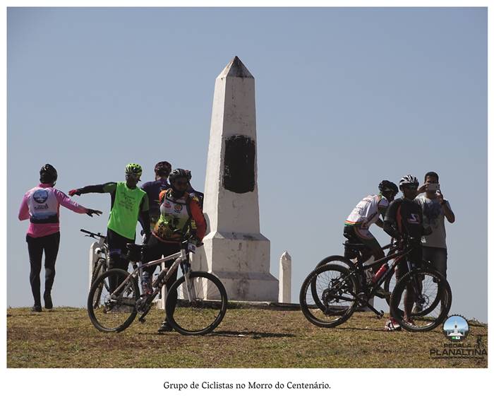 4 z 5: Grupo de Ciclistas no Morro do Centenário. Mulheres e homens trajando roupas para a prática do ciclismo e capacetes estão rodeando a Pedra Fundamental. Realização: Academia Planaltinense de Letras, Artes e Ciências (APLAC), pelo Ecomuseu Pedra Fundamental e pelo Coletivo Nativo Audiodescrição produzida  pelo Instituto de Promoção das Pessoas com Deficiência Visual Audiodescritora: Elma Lúcia Rodrigues Consultor: Fernando Rodrigues Este projeto é promovido com recursos do Fundo de Apoio a Cultura do DF